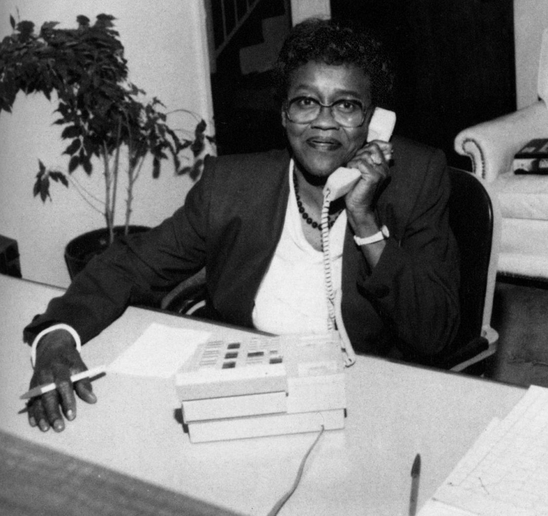 A woman wearing glasses and a suit jacket sits at a desk, smiling while talking on a corded phone. She holds a pen in one hand, with a stack of tapes and papers on the desk in front of her.