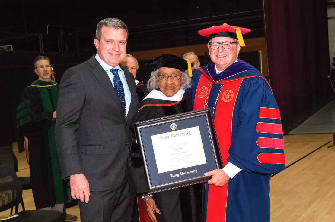 Three people pose at a graduation ceremony. One holds a framed diploma from King University. All wear academic regalia, and the group smiles for the photo on a gymnasium floor. Others stand in the background.