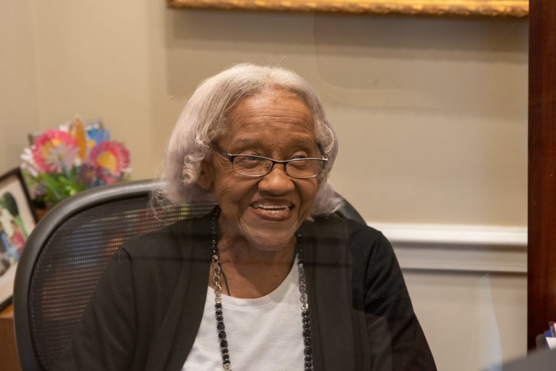 An elderly woman with gray hair and glasses sits in an office chair, smiling. She wears a black sweater, white blouse, and a beaded necklace. Colorful flowers and a photo are visible in the background on a desk.