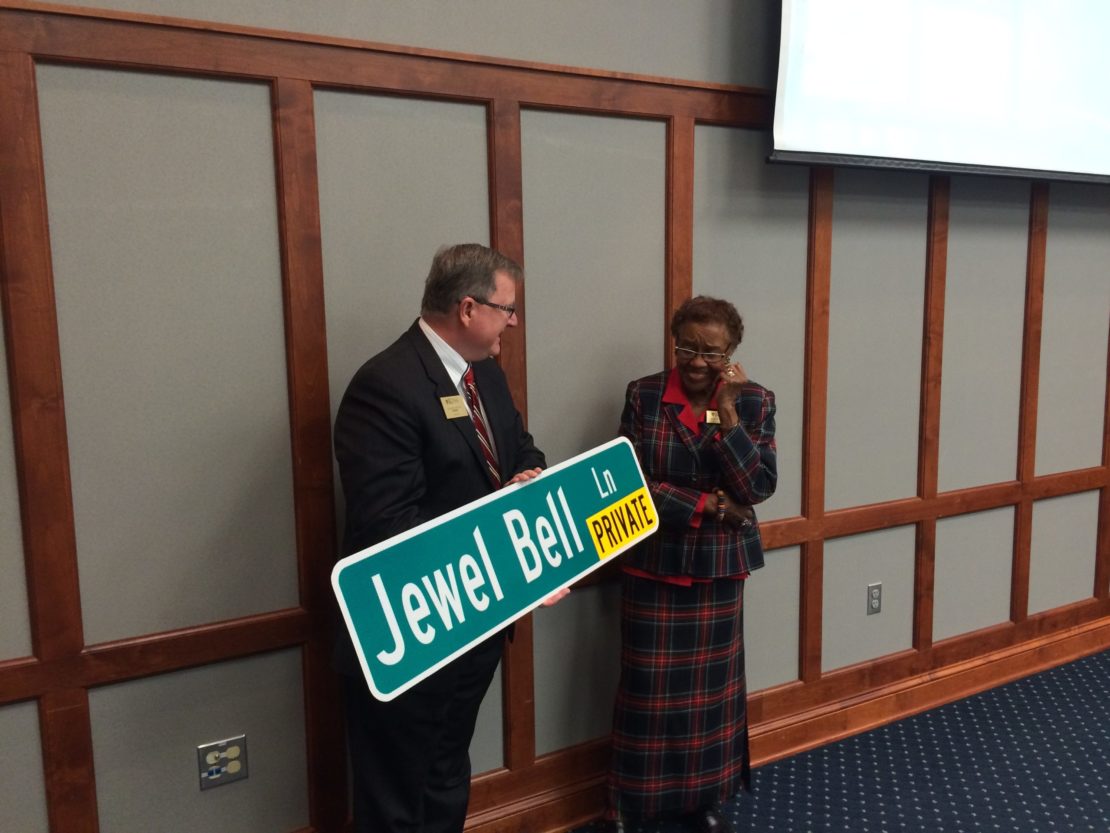 A man hands an older woman a green street sign reading Jewel Bell Ln PRIVATE in a formal room with wood paneling; both are smiling and dressed in business attire.