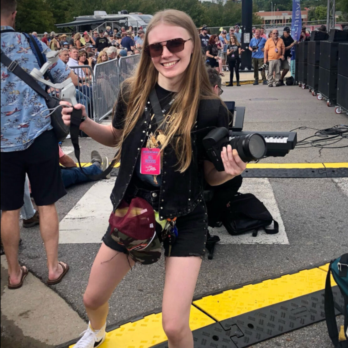A smiling young woman with long blonde hair, sunglasses, and a camera stands outdoors at an event. She wears a black outfit, black boots, and a pink staff badge, with a crowd and equipment in the background.