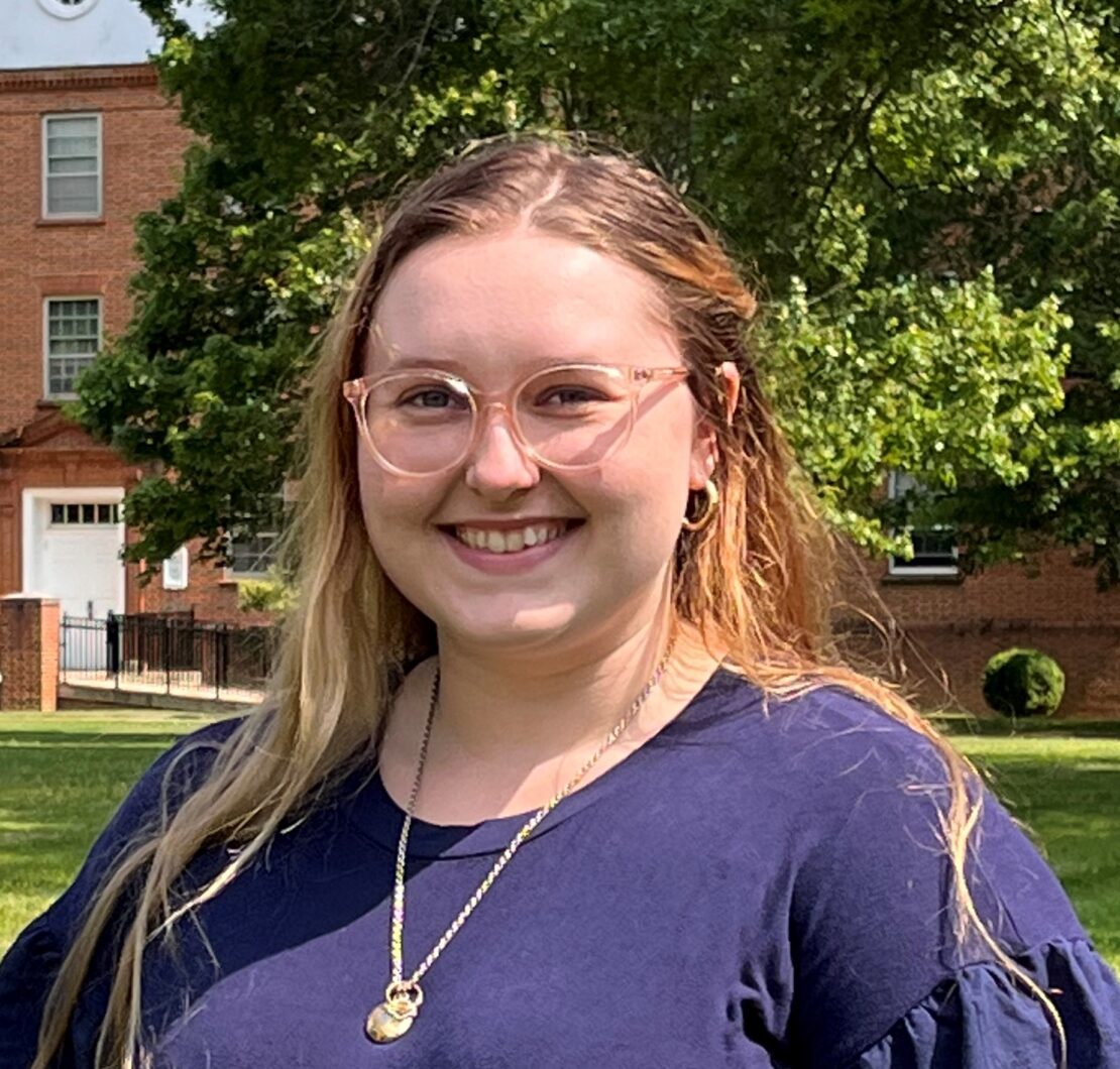 A young woman with long blonde hair and glasses smiles outdoors. She is wearing a navy blue top and gold necklace, standing in front of trees and a red brick building.