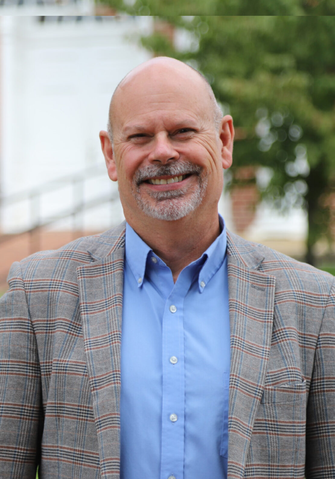 A smiling bald man with a gray goatee wearing a blue shirt and a plaid blazer stands outdoors in front of blurred greenery and a building.