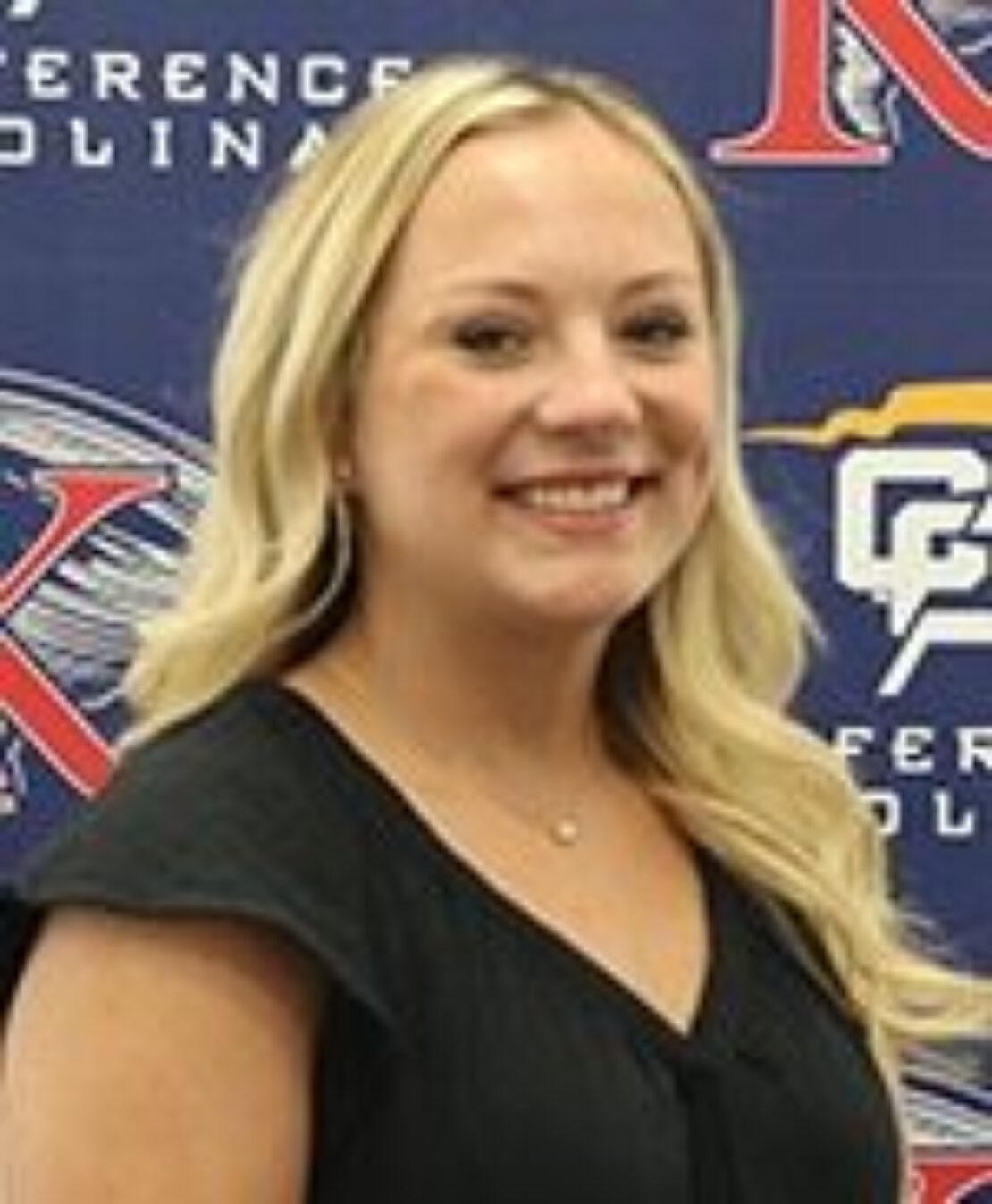 A smiling woman with long blonde hair wearing a black top stands in front of a blue and red conference banner.