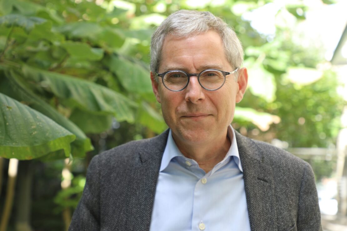 A middle-aged man with short gray hair and glasses, wearing a blue shirt and gray blazer, stands outdoors in front of lush green foliage, smiling slightly at the camera.