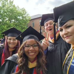 Four graduates in caps and gowns smile for a selfie outdoors. One person flashes a peace sign. Trees and a brick building are visible in the background.
