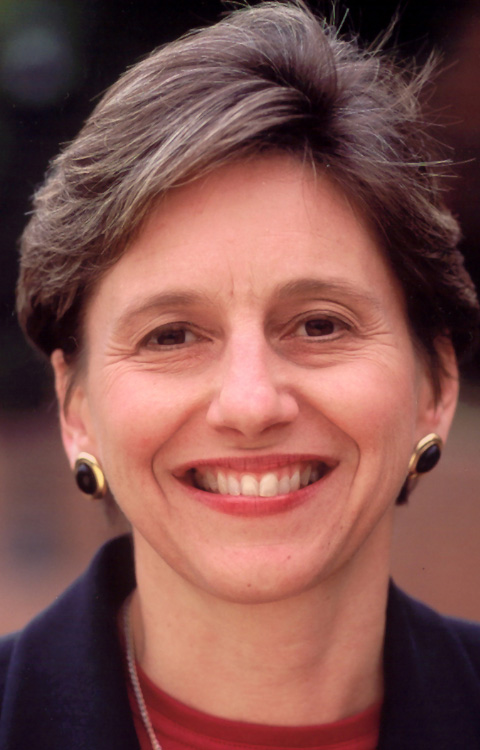 A woman with short brown hair smiles at the camera. She is wearing gold and black earrings, a dark jacket, and a red top. The background is softly blurred.