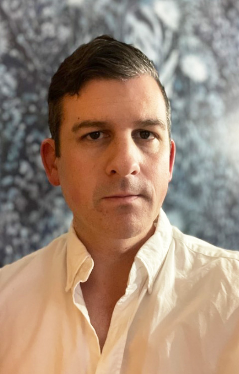 A man with short dark hair wearing a white button-up shirt looks at the camera with a neutral expression. The background is blurred with blue and white tones.