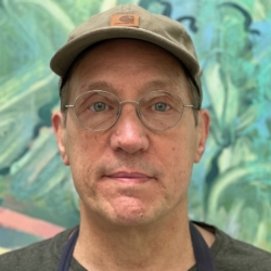 A middle-aged man wearing glasses, a gray cap, and a dark shirt stands in front of a colorful, abstract background. He looks directly at the camera with a neutral expression.