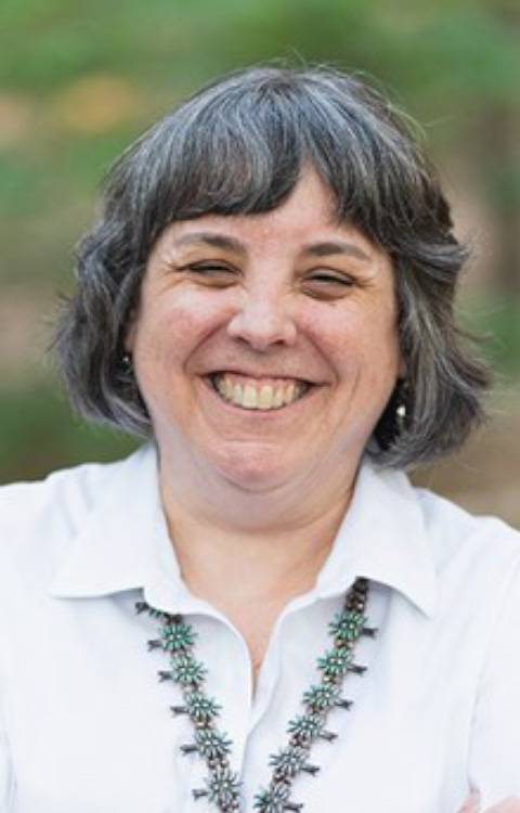 A middle-aged woman with short gray hair smiles broadly, wearing a white collared shirt and a turquoise necklace. The background is softly blurred greenery.