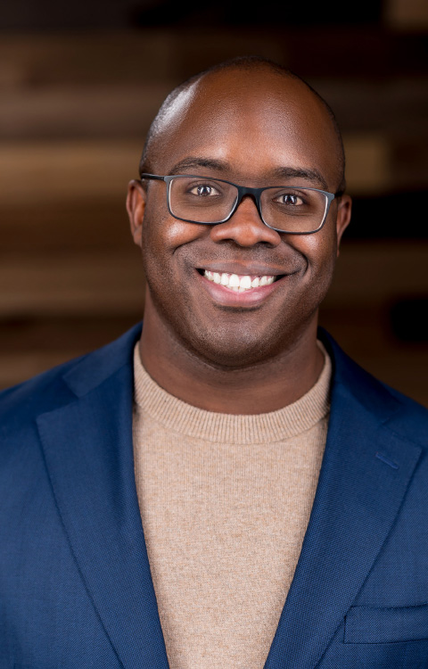 A man with glasses smiles at the camera. He is wearing a blue blazer over a beige sweater, and the background features blurred wooden panels.