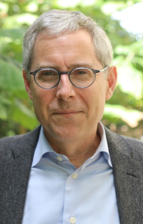 A middle-aged man with short gray hair and glasses is wearing a light blue shirt and a gray blazer, standing outdoors with green foliage in the background.