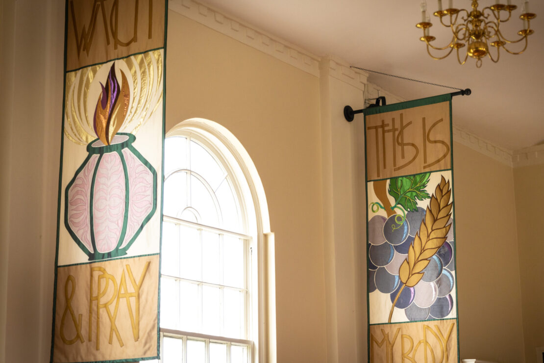 Two decorative fabric banners hang near a tall arched window; one reads WATCH & PRAY with a burning flame, and the other says THIS IS MY BODY with grapes and wheat. A gold chandelier hangs above.
