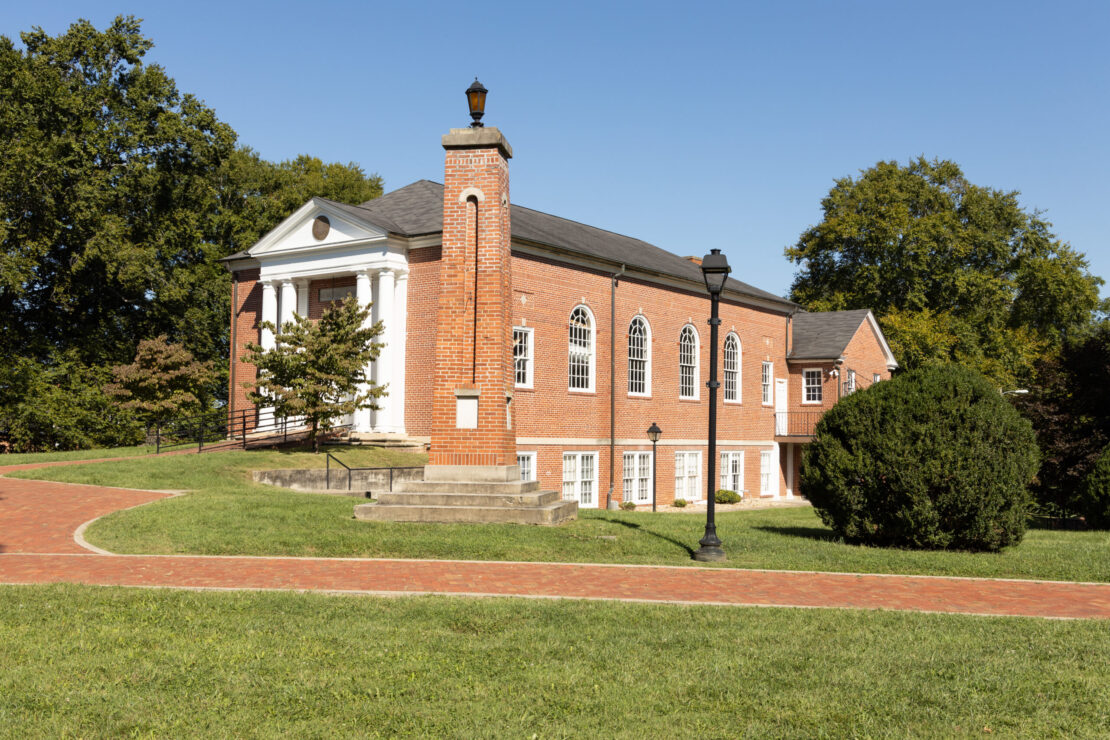 A red brick building with white columns and arched windows stands on a grassy lawn, reflecting the connection between faith and culture. A curved brick walkway, black lamp posts, and trees complete the scene under a clear blue sky.