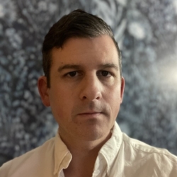 A man with short dark hair and a serious expression wears a white collared shirt, standing in front of a blurred, dark blue patterned background.