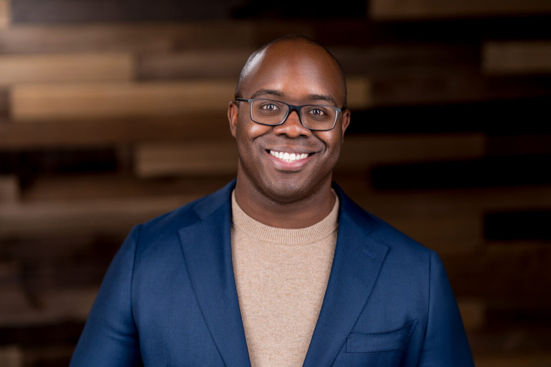 A smiling man wearing glasses, a beige sweater, and a blue blazer stands in front of a blurred wooden wall background.