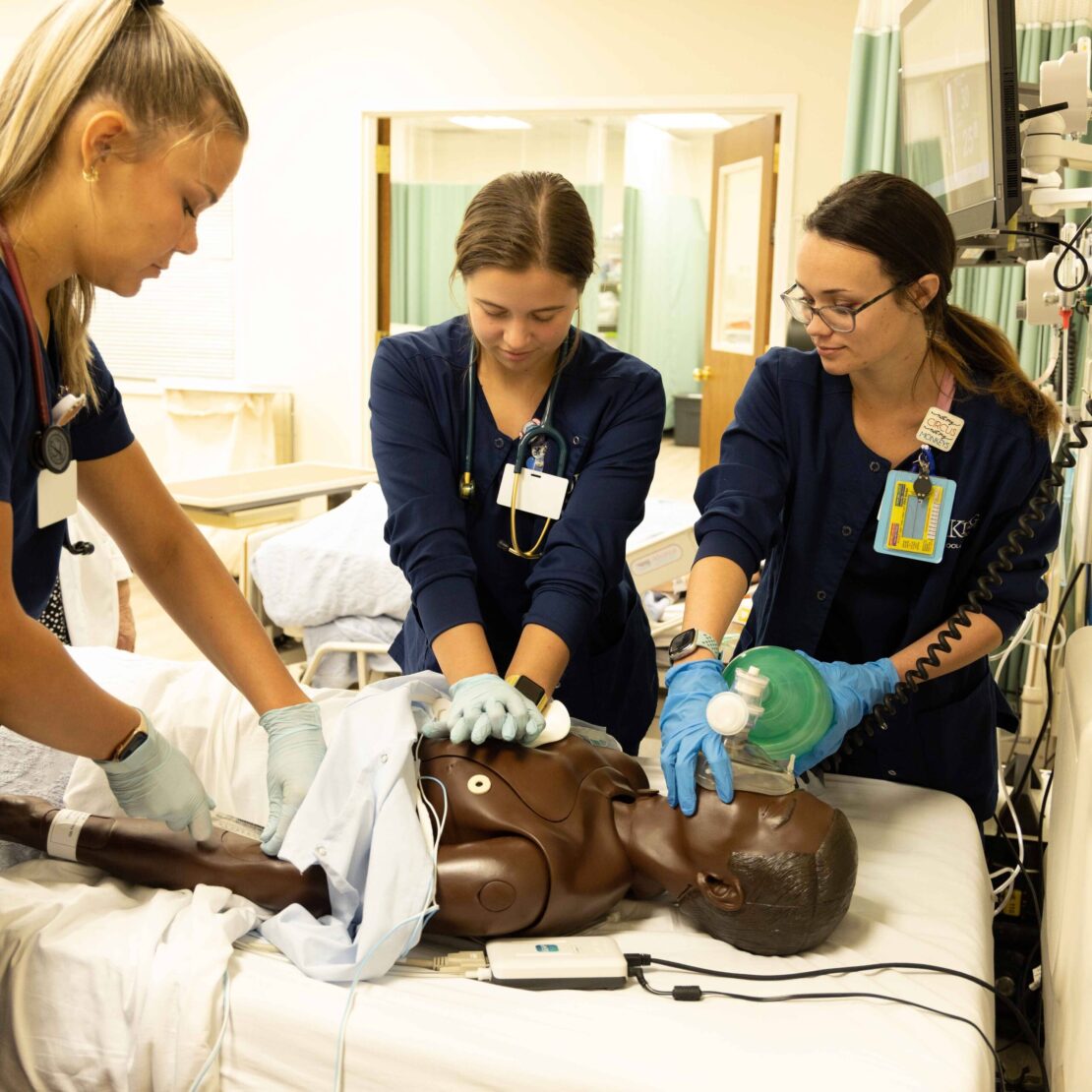 Three medical professionals practice CPR and ventilation on a medical mannequin in a clinical training room, using medical equipment and wearing gloves and scrubs.