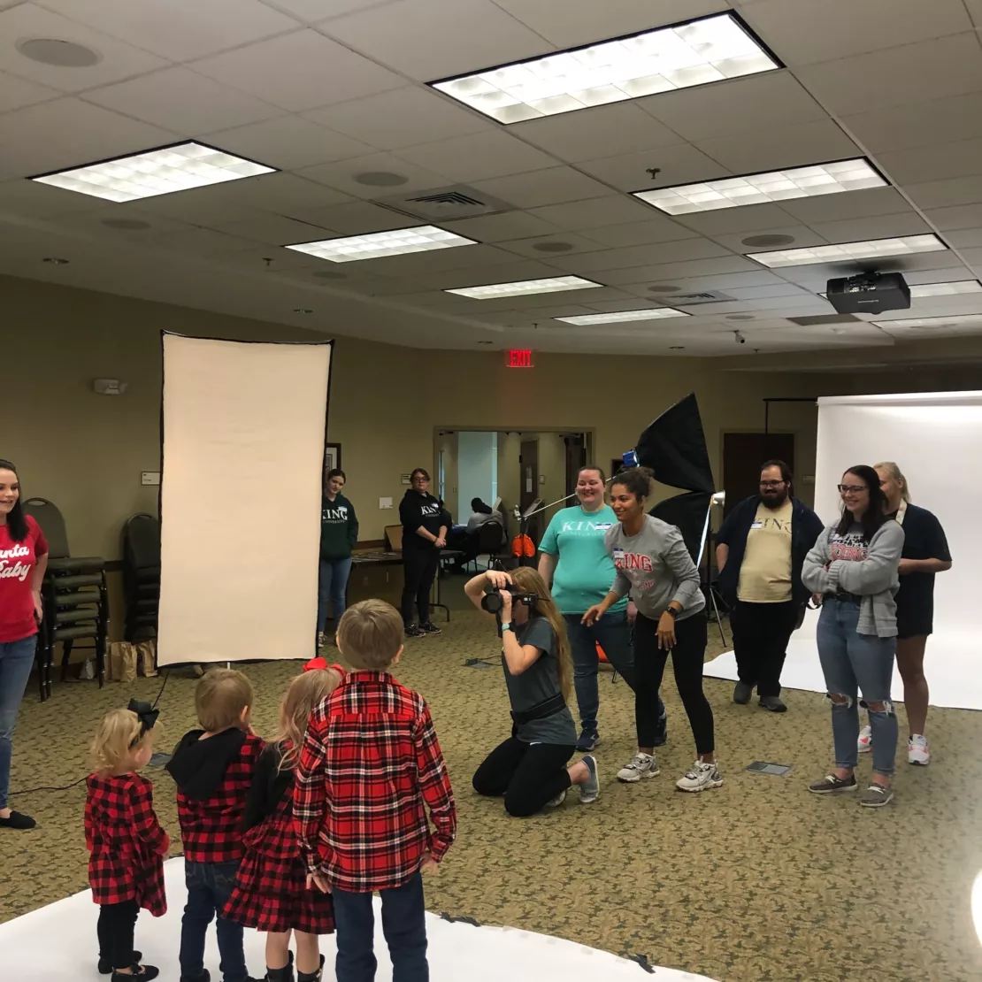 A group of children in matching red plaid outfits pose for a photo while several adults, including the photographer, stand around them in a brightly lit indoor studio with backdrops and lighting equipment.
