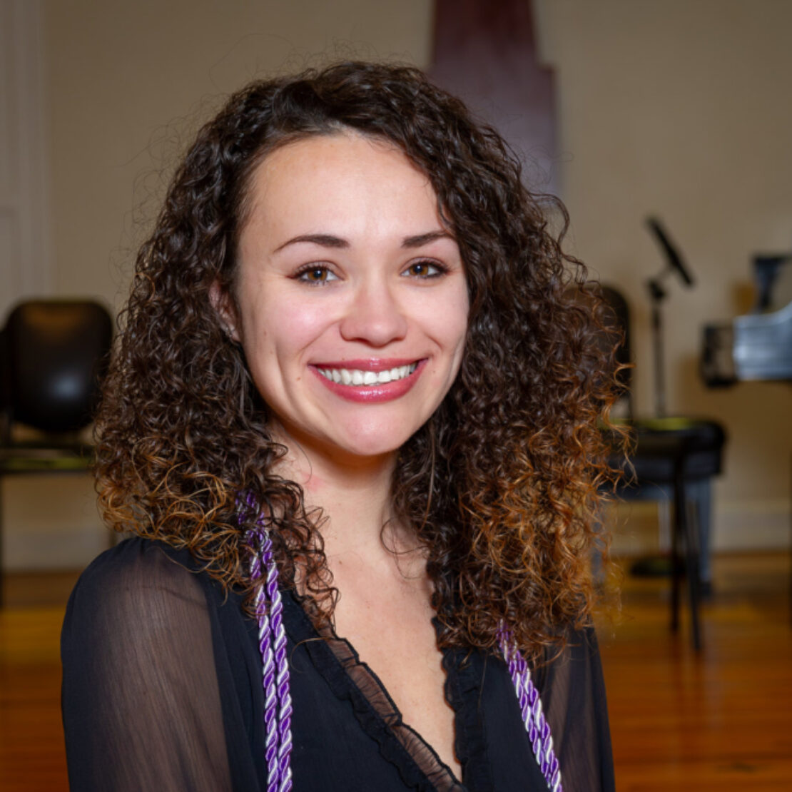 A young woman with curly brown hair wearing a black blouse and honor cords smiles at the camera. She is indoors, with chairs and a microphone stand visible in the background.
