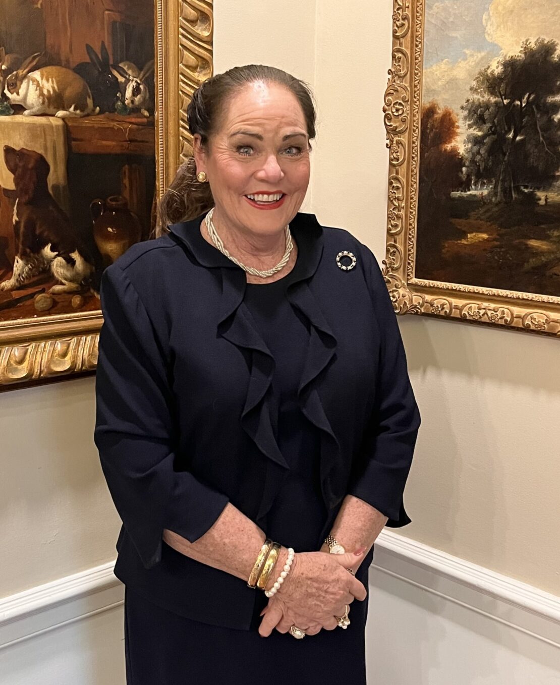 A woman with brown hair tied back, wearing a navy outfit with pearls and gold bracelets, stands smiling in front of framed paintings on a beige wall.