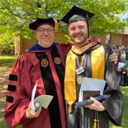 Two people in graduation attire smile and pose outdoors. One wears a maroon academic robe and cap, and the other wears a black cap and gown with a gold stole. Both hold diplomas. Green trees and a brick building are in the background.