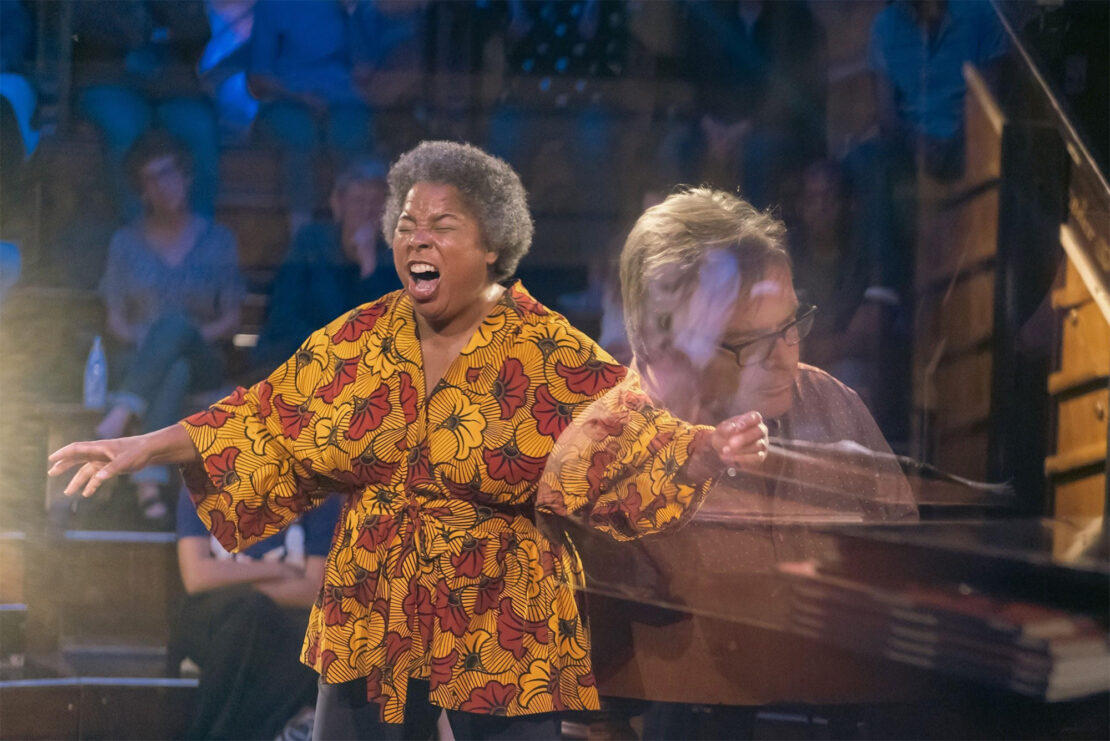 A woman passionately sings on stage in a yellow and red floral top while a pianist, seen through a glass reflection, plays beside her. Audience members watch in the dimly lit background.