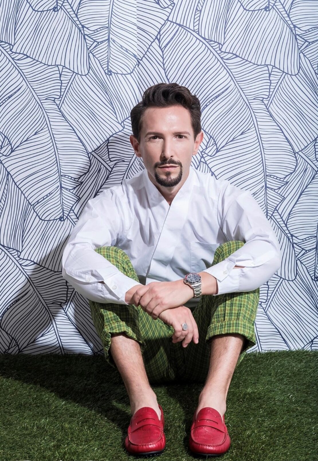 A man with short brown hair and a trimmed beard sits on artificial grass in front of a patterned white and black wall. He wears a white shirt, green plaid pants, a watch, and red loafers.
