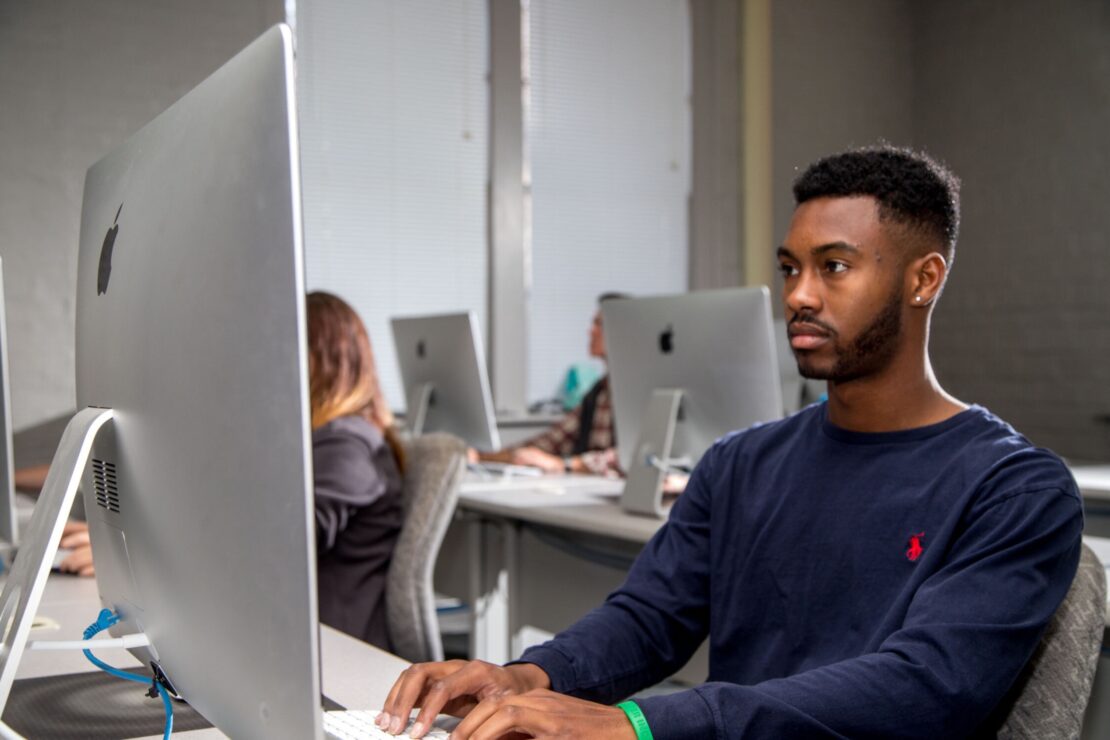 A young man sits at a desk using an Apple desktop computer in a classroom with other students working at similar computers in the background.
