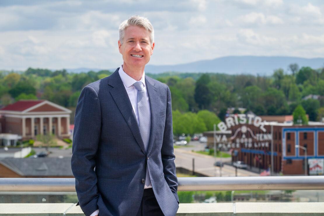 A man in a gray suit and tie stands smiling on a rooftop with a scenic view of Bristol, Virginia, including the city sign and distant mountains, under a partly cloudy sky.