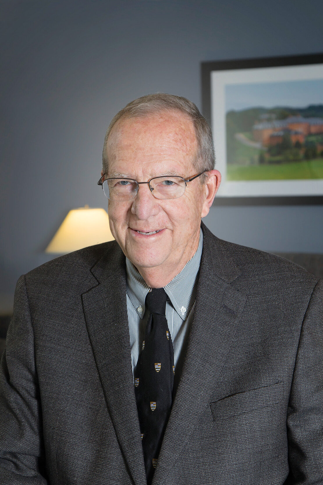 An older man with glasses wearing a brown suit jacket, striped shirt, and black tie smiles at the camera; a lamp and a framed landscape photo are in the background.