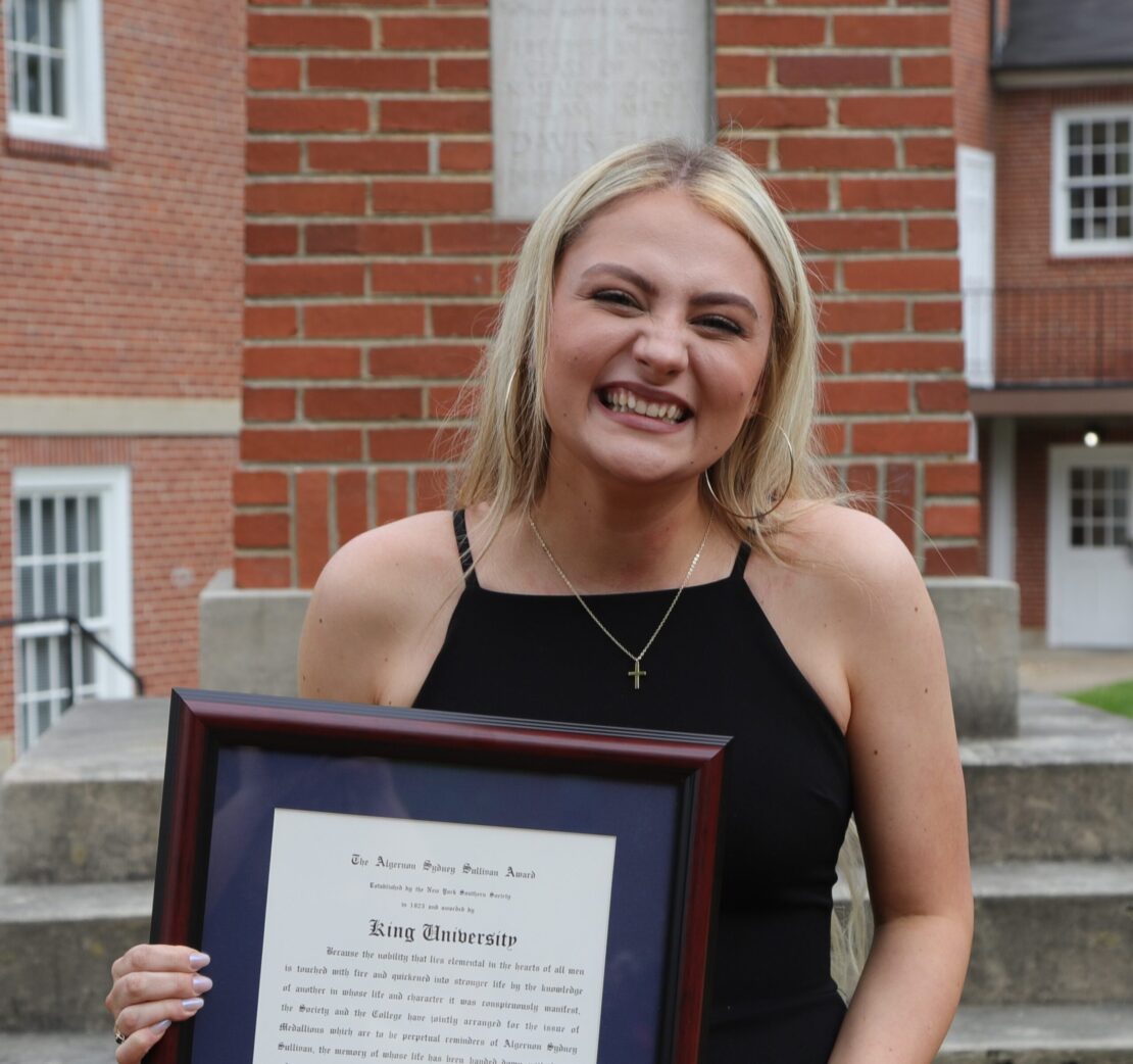 A young woman with long blonde hair smiles broadly while holding a framed certificate. She stands outside in front of brick buildings and steps, wearing a black dress and a necklace with a cross pendant.