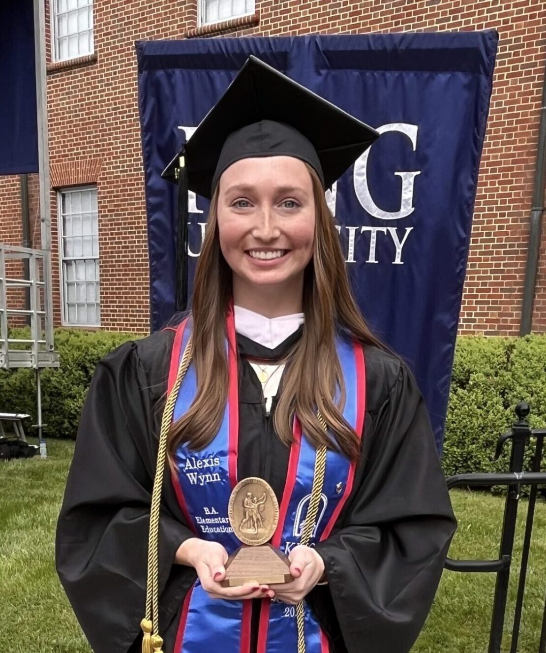 A young woman in a graduation cap and gown smiles while holding an award. She stands outdoors in front of a building and a large blue university banner.
