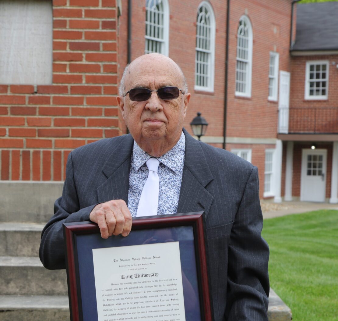 An older man in a suit, tie, and sunglasses stands outdoors holding a framed certificate. Behind him is a red brick building with white-trimmed windows and a staircase to the side.