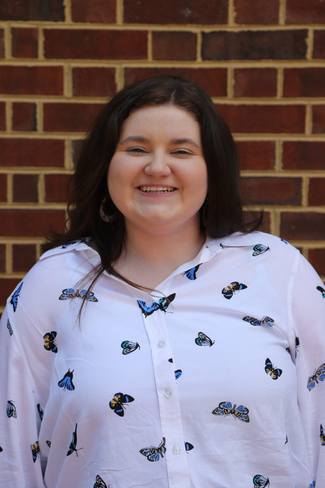 A woman with long brown hair, wearing a white shirt with a butterfly print, stands smiling in front of a brick wall.