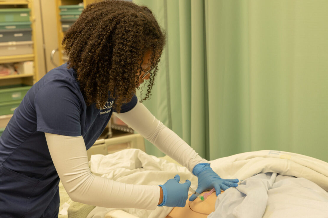 A healthcare worker in blue scrubs and gloves practices a medical procedure on a mannequin in a clinical training room.