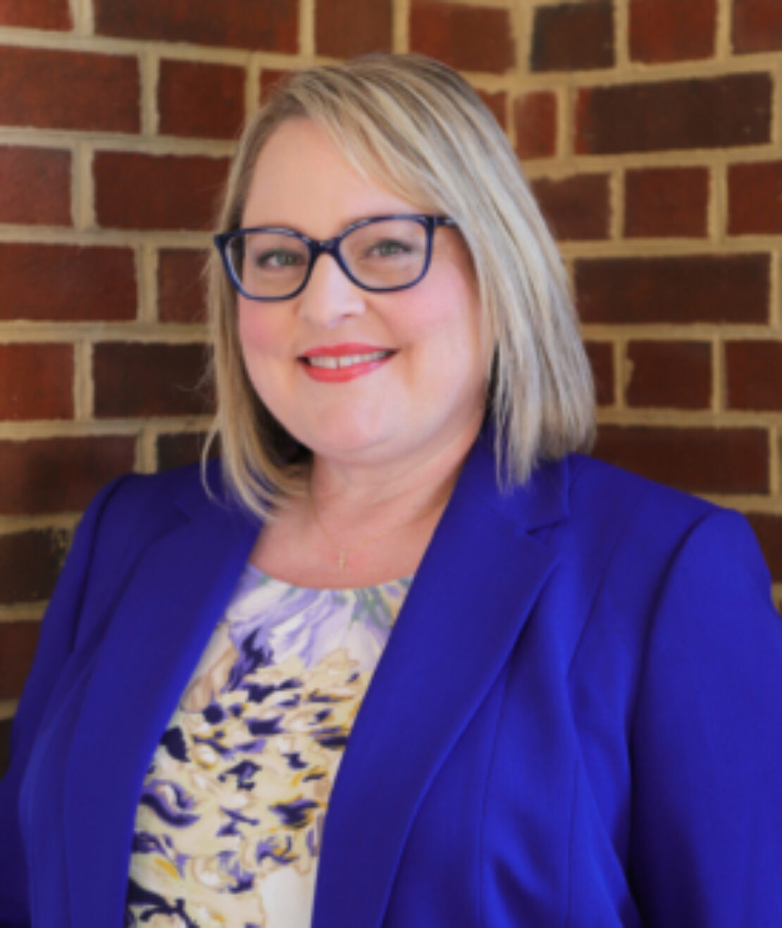 A woman with shoulder-length blonde hair, wearing glasses, a blue blazer, and a floral top, smiles while standing in front of a brick wall.