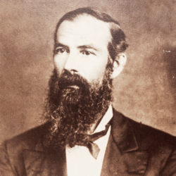 A sepia-toned portrait of a man with a long, full beard and mustache, wearing a suit, white shirt, and bow tie, looking slightly to the side against a plain background.