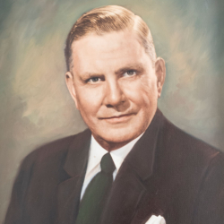 A formal portrait of a middle-aged man with short, slicked-back blonde hair, wearing a dark suit, white shirt, and dark tie, set against a softly blurred, neutral background.
