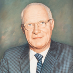 A middle-aged man with short white hair and glasses, wearing a blue checkered suit, white shirt, and striped tie, poses against a soft, muted green background.
