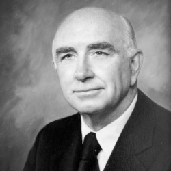 Black and white portrait of an older man with a bald head wearing a dark suit, white shirt, and dark tie, looking directly at the camera with a neutral expression.