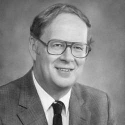 A middle-aged man with glasses and short hair, wearing a suit, dress shirt, and tie, smiling at the camera in a black and white portrait.