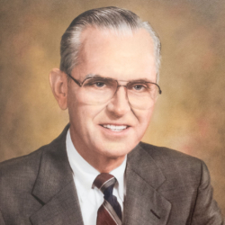 Portrait of an older man with gray hair, wearing glasses, a brown suit, white shirt, and striped tie, smiling against a softly blurred, warm-toned background.