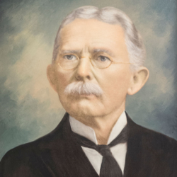 Portrait of an older man with gray hair, a mustache, wire-rimmed glasses, and a suit with a bow tie, posed against a soft, neutral background.