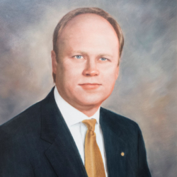 A formal portrait of a man in a dark suit, white shirt, and gold tie, posing against a soft, blurred background. He has short, light hair and a neutral expression.