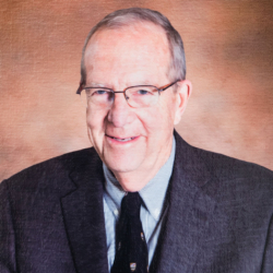 An older man with short gray hair and glasses, wearing a dark suit, light blue shirt, and patterned tie, smiles in front of a warm brown gradient background.