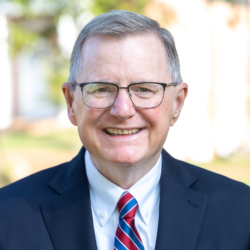 Smiling older man with short gray hair, wearing glasses, a dark suit, white shirt, and a red and blue striped tie. The background is blurred greenery and light-colored buildings.