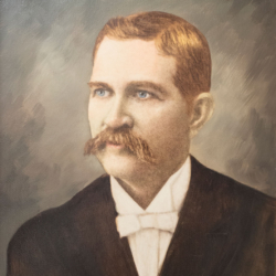 A sepia-toned portrait of a man with light brown hair, a thick mustache, blue eyes, and fair skin, wearing a dark suit, white shirt, and white bow tie, against a muted background.