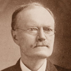 Sepia-toned portrait of an older man with glasses, a mustache, and short hair, wearing a suit and tie, looking slightly to the side.