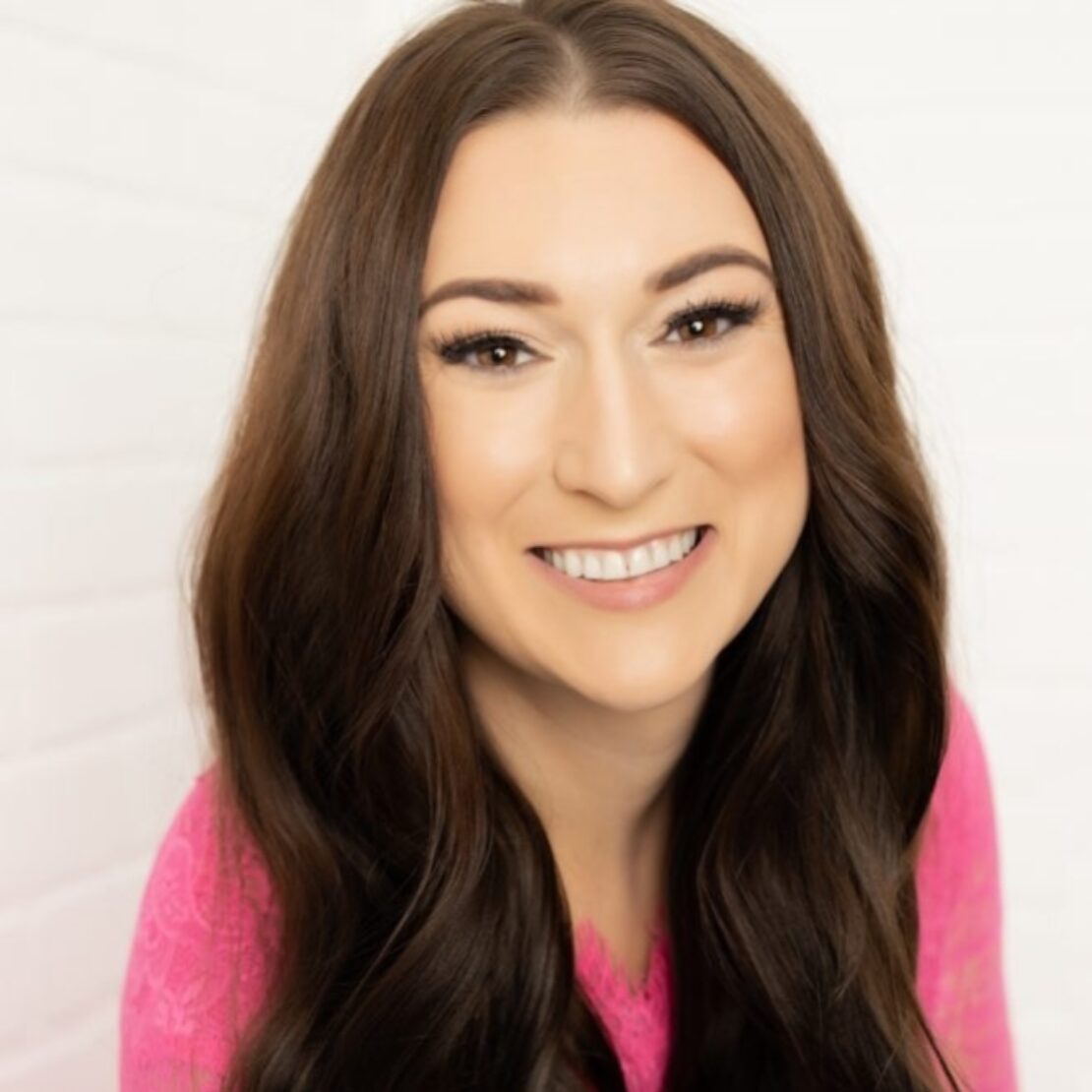 Smiling woman with long brown hair wearing a bright pink top, posing in front of a white brick wall background.