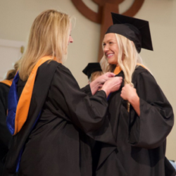 Two people in graduation gowns and caps are smiling at each other. One person is placing a graduation hood over the other’s shoulders during a graduation ceremony.